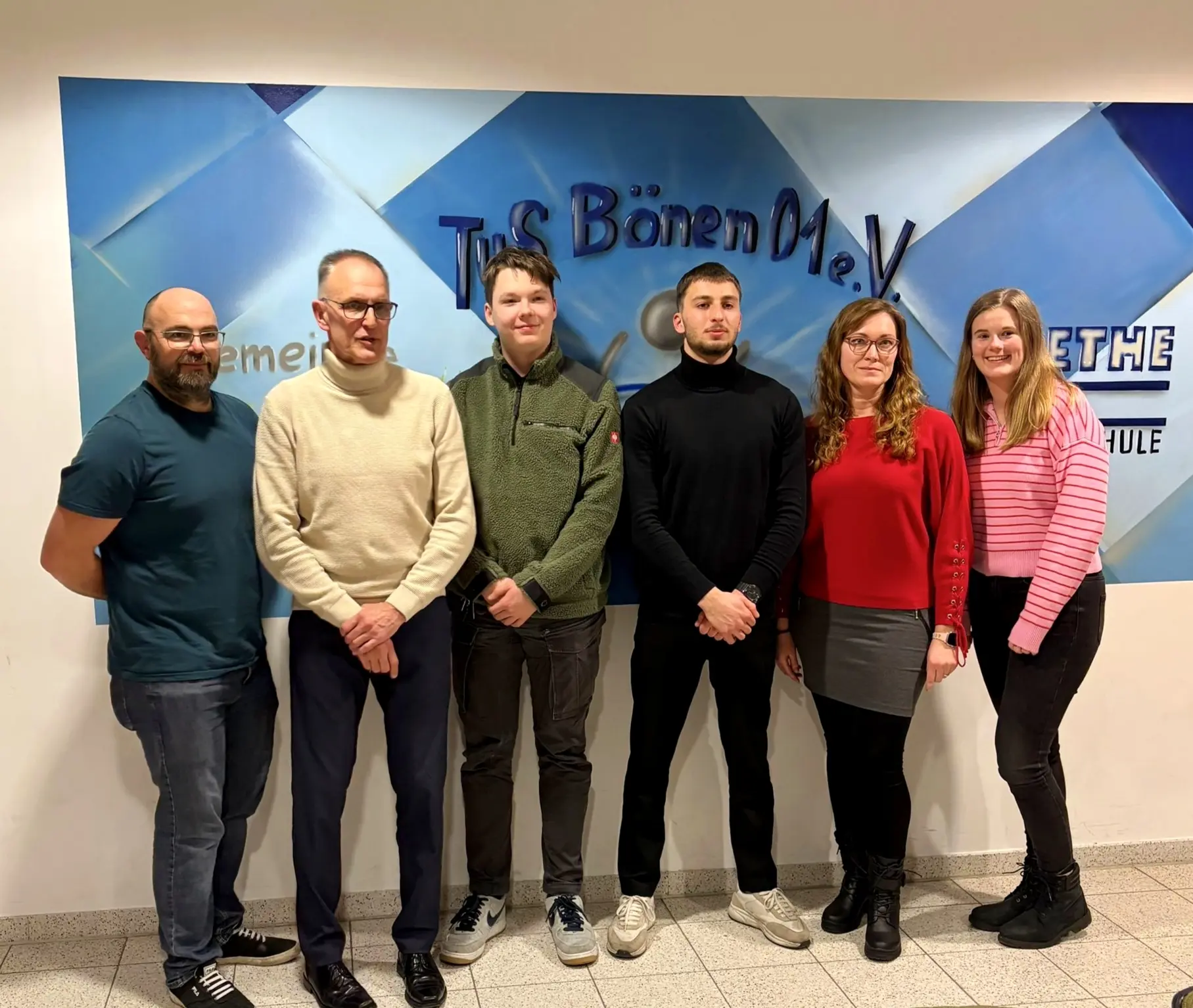 Sechs Personen stehen vor einer Wand mit dem Logo von TuS Bönen e.V. in einem Innenraum. Die Gruppe besteht aus drei Männern und drei Frauen.