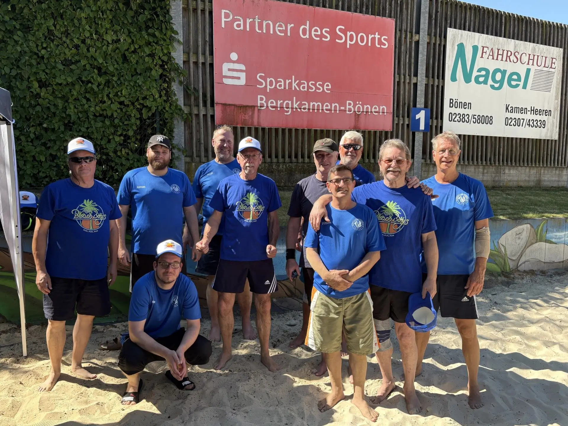 Mehrere Männer in blauen Trikots stehen und hocken auf dem Sandplatz vor Sponsortafeln beim Beachvolleyballturnier in Bönen unter freiem Himmel.